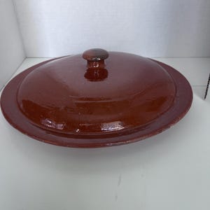 May include: A reddish-brown ceramic casserole dish with a lid. The lid has a rounded shape with a small, round handle. The dish has a glossy finish and a slightly raised rim. The overall design is simple and functional.