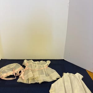 May include: A set of doll clothing consisting of a sheer off-white dress with lace trim and a pink ribbon tie, a matching bonnet with lace trim and pink ribbon ties, and a separate sleeveless off-white dress with a lace-trimmed hemline.