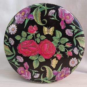 May include: Round black decorative tin with a floral design. The tin features pink roses, purple irises, white daisies, and yellow butterflies. Green leaves and stems are interspersed throughout the design.