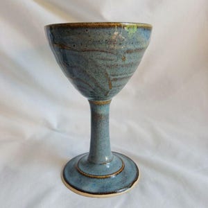 May include: A handmade ceramic goblet with a wide bowl and a slender stem. The goblet is glazed in shades of blue and brown, with a textured surface. The base is round and the rim is a darker brown. The goblet is approximately 18 cm tall.