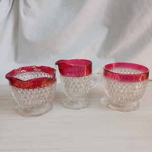 Puede incluir: Un juego de crema y azucarero de cristal vintage. El juego incluye una jarra de crema con asa, un azucarero y un cuenco pequeño. Cada pieza tiene un patrón transparente tallado en diamante con un borde rojo vibrante.