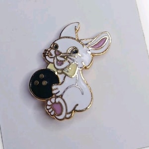 以下が含まれることがあります： 金色の縁取りが付いた白いウサギのピンバッジ。黒いボウリングボールを持っています。ウサギはピンク色の内側の耳と足、黄色の蝶ネクタイをしており、笑顔です。白い背景にあります。