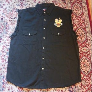 Pode incluir: Camisa preta sem mangas com botões, dois bolsos no peito e gola. A camisa apresenta um patch bordado colorido com chamas e texto. Os ombros têm uma borda desfiada.