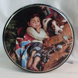 Pode incluir: Lata decorativa redonda com uma pintura de uma criança a dormir e um cão. A criança está deitada numa colcha com um camião de brinquedo e outros brinquedos espalhados. O cão é castanho e branco. A lata tem uma borda preta.