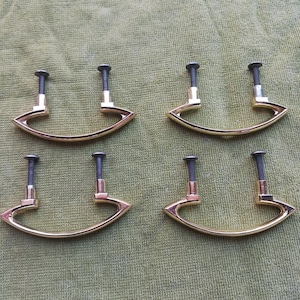 May include: Four gold-toned cabinet pulls with a curved handle design. Each pull has a dark gray screw for mounting. The pulls are arranged on a green fabric surface.