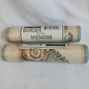 May include: Two rolls of wallpaper border with a seashell and plant design. The rolls are wrapped in clear plastic. The text on the label reads "BORDER" and "SKB34059B". The border has a beige background with green and brown accents.