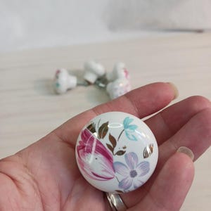May include: White ceramic cabinet knobs with floral designs in pink, blue, and gold. The knobs are round with a glossy finish. Several knobs are visible, suggesting a set for furniture or decor.