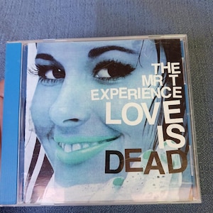 May include: A compact disc case featuring a blue-tinted close-up of a face with light green lips. The text on the cover reads THE MR T EXPERIENCE LOVE IS DEAD in white and black lettering. The case has a bright blue spine and clear plastic front.