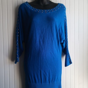 May include: A vibrant blue sweater dress with a boat neck and three-quarter sleeves. The neckline and sleeves are embellished with small, silver-colored studs. The dress is displayed on a black mannequin against a white-paneled background.