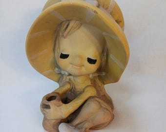 Könnte beinhalten: Keramikfigur eines sitzenden Kindes mit einem großen gelben Hut mit weißen Streifen. Das Kind hält einen kleinen Topf und hat geschlossene Augen mit schwarzen Wimpern. Die Figur ist in Gelb-, Beige- und Brauntönen bemalt.