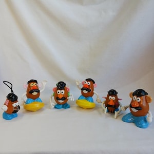 May include: A collection of six Mr. Potato Head toy figures. The figures have brown potato-shaped heads, black hats, and mustaches. They are wearing blue and yellow clothing and have white arms and hands. One figure has a black string attached.