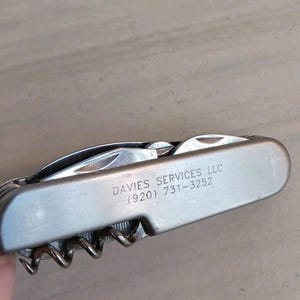 Op de afbeelding: Een zilverkleurig multi-tool met een kurkentrekker, mes en andere gereedschappen. De tekst "DAVIES SERVICES LLC (920) 731-3252" staat op de zijkant. De tool is gedeeltelijk open, waardoor enkele onderdelen zichtbaar zijn.