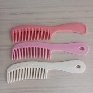May include: Three plastic combs in shades of pink and white. The top comb is a solid pink, the middle comb is light pink with the word "Unbreakable" printed on it, and the bottom comb is white with the same text.