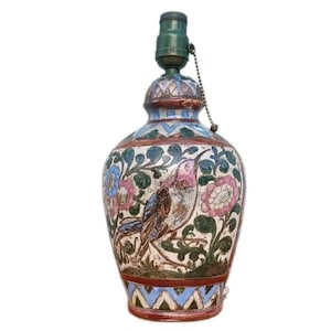 Könnte beinhalten: Dekorative Keramiklampe mit einem bemalten Vogel- und Blumendesign. Die Lampe hat einen grünen Aufsatz, eine Zugkette und einen Sockel mit blauen, roten und weißen geometrischen Mustern. Der Korpus der Lampe ist mit rosa, blauen und grünen Blumen- und Vogelmotiven verziert.