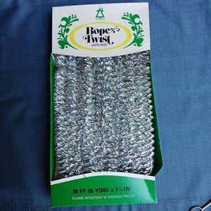 Pode incluir: Um pacote de guirlanda prateada Rope Twist. O cabeçalho branco apresenta adornos decorativos verdes e o texto Rope Twist GARLAND. A base verde exibe o texto 18 FT (6 YDS) x 1 1/2 IN, FLAME RESISTANT & TARNISH PROOF.