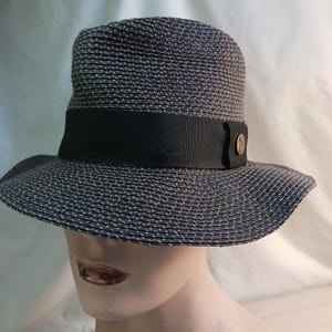 May include: A blue and tan woven hat with a black band and a decorative button. The hat has a wide brim and a cylindrical crown. The hat is designed to be worn outdoors.