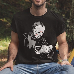Könnte beinhalten: Ein Mann trägt ein schwarzes T-Shirt mit einem Aufdruck eines Hockeymasken-tragenden Mannes, der einen weißen Clog hält. Das T-Shirt trägt den Text "Freitag der 13.". Der Mann sitzt auf einem Baumstamm in einer Waldlandschaft.