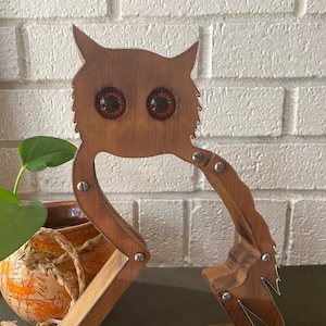 Puede incluir: Una escultura de búho de madera con grandes ojos redondos de color naranja. El búho está hecho de madera y tiene un acabado rústico y desgastado. El búho está de pie sobre una base triangular.
