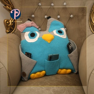 May include: A handmade crochet owl-shaped pillow in shades of blue, gray, white, and yellow. The pillow has pockets for remote controls and a phone. A pink bow is attached to the top of the owl's head. The pillow is placed on a beige armchair.
