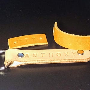 May include: Two leather keychains with metal rings. One keychain is tan and has the name "ANTHONY" embossed on it. The other keychain is brown and has the number "777" embossed on it.