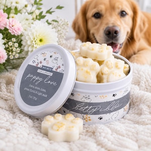 May include: A round white metal tin filled with off-white paw-shaped wax melts. The lid and side label feature the text "puppy love", "odor eliminator", "betty blu", and "3oz | 85g", along with "lemon, ozone, linen, green floral & light musk".