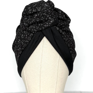 May include: A black turban featuring a floral design in grey. The turban is made from a soft material and has a twisted knot detail at the top. The base of the turban is plain black.