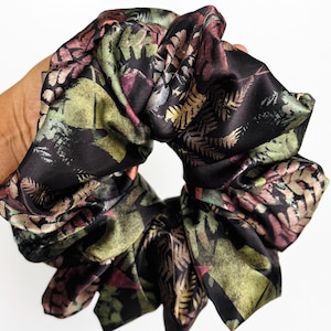 May include: A large, black scrunchie with a botanical print featuring green, gold, and burgundy leaves. The fabric has a silky sheen, and the scrunchie is gathered and voluminous, suitable for various hairstyles.