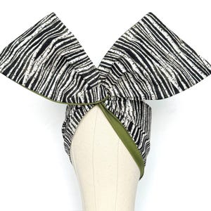 May include: A turban-style head wrap featuring a black and white zebra stripe design. The edges are trimmed with green fabric. The head wrap is displayed on a mannequin head.