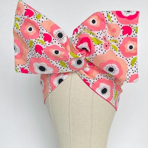 May include: A pink and white headband with a floral pattern. The headband is tied in a bow on top of the head.