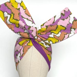 May include: A vibrant turban-style head wrap with a large bow. The fabric displays an abstract pattern in yellow, pink, purple, and orange. A solid purple band accents the base, creating a fashionable accessory.