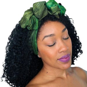 May include: A green headband with a textured pattern is worn on a person's head. The headband is made of a soft material and has a large, decorative knot on top. The person has dark curly hair and is wearing purple lipstick.