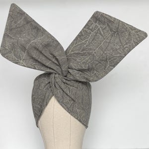 May include: A grey turban-style headband with a knotted front and two large, upward-sweeping fabric pieces. The fabric has a subtle, textured pattern resembling leaf veins. The headband is displayed on a mannequin head against a white background.
