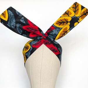 May include: A patterned headband with a knotted design, showcasing a mix of red leaves and yellow floral motifs against a black background. The headband is displayed on a mannequin head. This hair accessory is a stylish addition to any outfit.