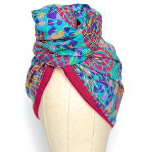 Peut inclure: Un turban coloré avec un motif floral vif en turquoise, violet, rose et or. Le turban a une bande magenta foncé à la base. Le tissu est enroulé et noué en haut, créant un couvre-chef élégant.