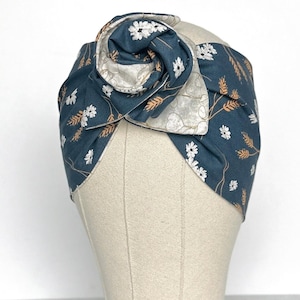 May include: A dark teal fabric headband featuring a floral pattern with white flowers and tan wheat stalks. The headband has a large, twisted knot detail at the centre front, showcasing a contrasting light grey fabric with a subtle leaf print.
