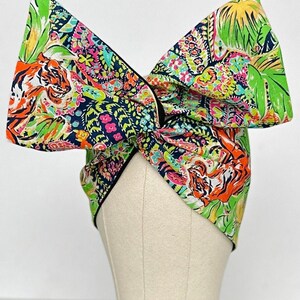 May include: A large, colourful bow headband with a paisley and tiger print. The headband is made of fabric and has a black trim.