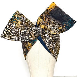 May include: A fabric headband featuring a large, upright bow with a mosaic-like pattern in shades of gold, black, and teal. The design consists of small, interlocking geometric shapes that create a textured visual effect across the entire surface.