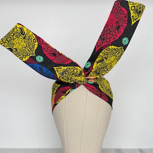 May include: A patterned headband with a knotted design. The fabric displays a repeating pattern of yellow, red, and blue leaf-like shapes on a black background. The headband is displayed on a mannequin head.