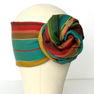 Puede incluir: Una colorida diadema con una gran flor decorativa. La diadema presenta rayas horizontales en turquesa, rojo, marrón y amarillo. La flor está hecha de la misma tela, con un diseño en espiral. La diadema se muestra en una cabeza de maniquí.
