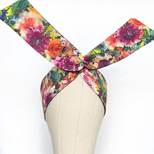 May include: A floral print headband with a knotted top and long, flowing ends. The headband features a vibrant watercolour design with pink, orange, and green flowers on a black background. The headband is displayed on a mannequin head.