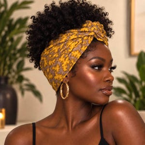May include: A wide, knotted fabric headband featuring a mustard yellow and grey leaf pattern. The headband is worn over dark, curly hair and is paired with large, circular gold hoop earrings. The fabric has a textured, matte appearance.