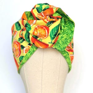 May include: A turban-style headband with a bright orange and green citrus fruit design on a yellow base. The fabric headband has a knotted detail at the top. The reverse side features a green leaf pattern. The headband is likely made of fabric.
