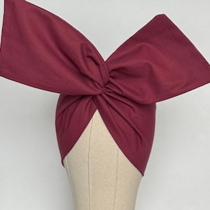 May include: A burgundy turban-style headband featuring a large, knotted bow. The fabric is a solid colour. The headband is displayed on a mannequin head. This accessory is suitable for various occasions and is a stylish head covering.