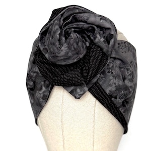 May include: A grey turban-style headband with a large fabric flower. The headband has a floral pattern and a textured black band. The flower is made from the same fabric as the headband, creating a cohesive design.