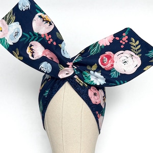 May include: A navy blue fabric headband featuring a large, upright bow detail. The fabric is patterned with a floral design in shades of light pink, white, and teal green, accented with small red berries and dark green leaves throughout the print.