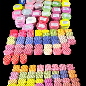 May include: A display of colourful bath sponges and soap bars. The sponges are in shades of pink, yellow, blue, and orange, with a waffle texture. The soap bars are oval-shaped and wrapped in white paper with labels.
