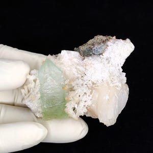 Könnte beinhalten: Ein Mineralexemplar mit einer markanten, durchscheinenden blassgrünen Kristallspitze, die an einer Ansammlung weißer, faseriger und kristalliner Formationen befestigt ist. Ein kleines, dunkles, strukturiertes graues Gesteinsfragment ist oben in der weißen Ansammlung eingebettet.
