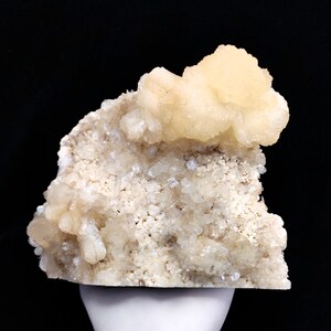 Pode incluir: Uma amostra mineral com um aglomerado de cristais amarelo-cremosos sobre uma matriz de cristais granulares menores. O mineral assenta sobre um pedestal branco contra um fundo preto, realçando a sua textura natural e variações de cor.