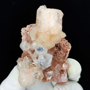 Könnte beinhalten: Ein Mineralexemplar mit einer Ansammlung durchscheinender, blass pfirsichfarbener Kristalle, die auf einer rauen, rotbraunen Matrix wachsen. Die Formation umfasst mehrere scharfe, kantige Kristallspitzen mit unterschiedlicher Klarheit.