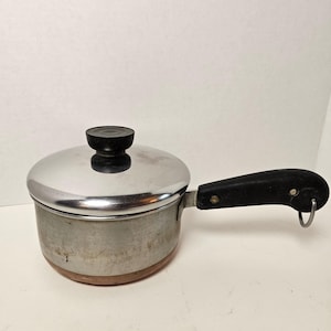 May include: A small, vintage saucepan with a lid. The pot is made of stainless steel with a copper-coloured base. It has a black handle and a black knob on the lid. The saucepan is designed for cooking.